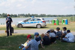 policjantka podczas pogadanki na dworze, mówi do dzieci w tle radiowóz i jezioro