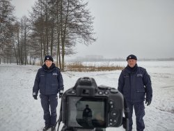 policjantka i policjant na tle jeziora, przed nimi aparat fotograficzny na statywie