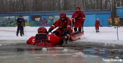 strażacy na zamarzniętym  jeziorze na saniach lodowych wyciągają z wody strażaka, który udaje osobę pod którą załamał się lód, wszyscy  ubrani w specjalne kombinezony do działań na lodzie i wodzie zimą, 
 w tle policjantka nagrywająca scenę aparatem