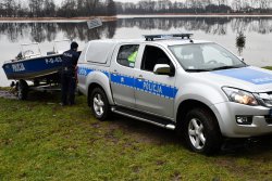 policyjna łódź podczas wodowania nad jeziorem, radiowóz wraz z wózkiem, na którym znajduje się łódka cofa w stronę jeziora, przy pojazdach stoi policjant